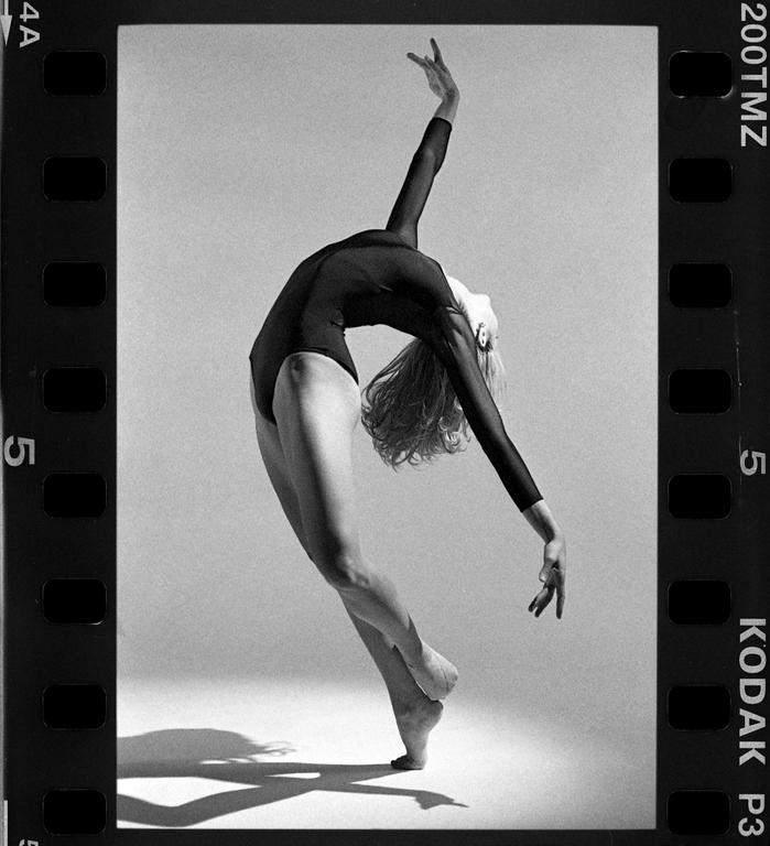A woman in a leotard is dancing with her back arched and one arm raised.