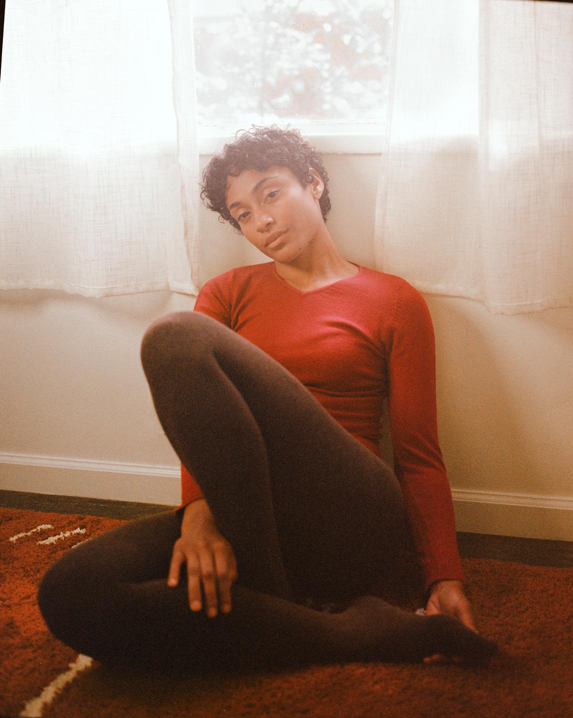 A woman with curly hair sits on a red rug next to a window with sheer white curtains.
