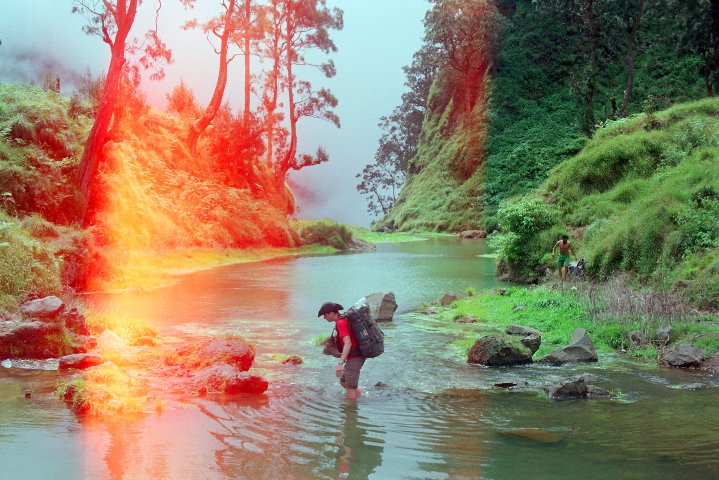 A man with a large backpack wades through a river with another man on the bank and a bright orange light leak on the left.