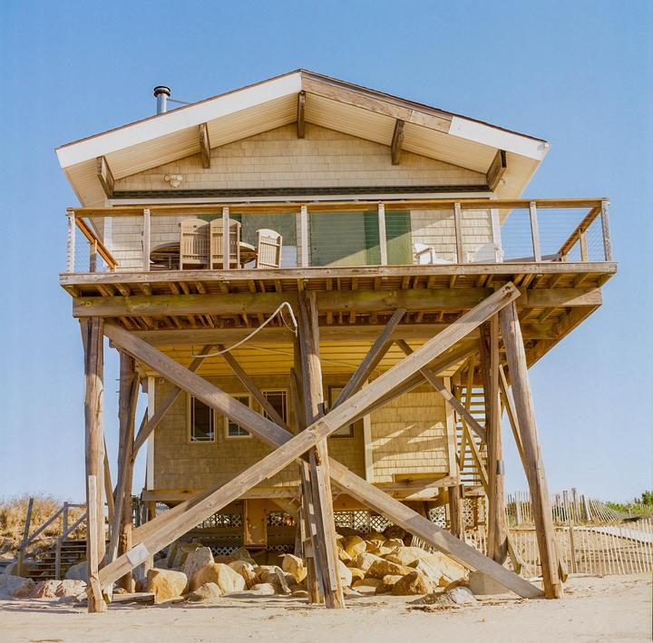A beach house on stilts stands on a sandy shore under a clear blue sky.