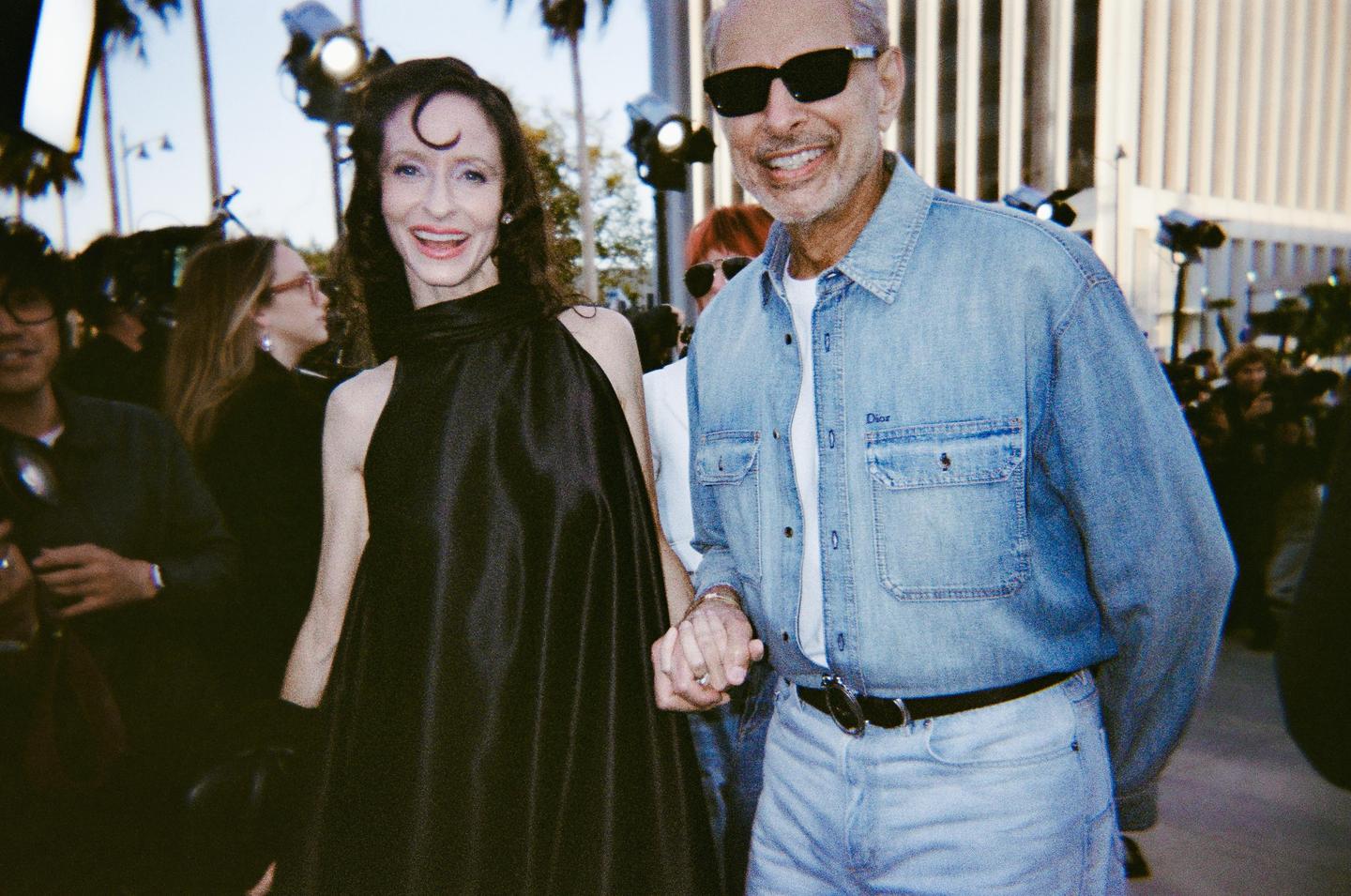 A couple smiles while holding hands on a red carpet with palm trees and lights in the background.