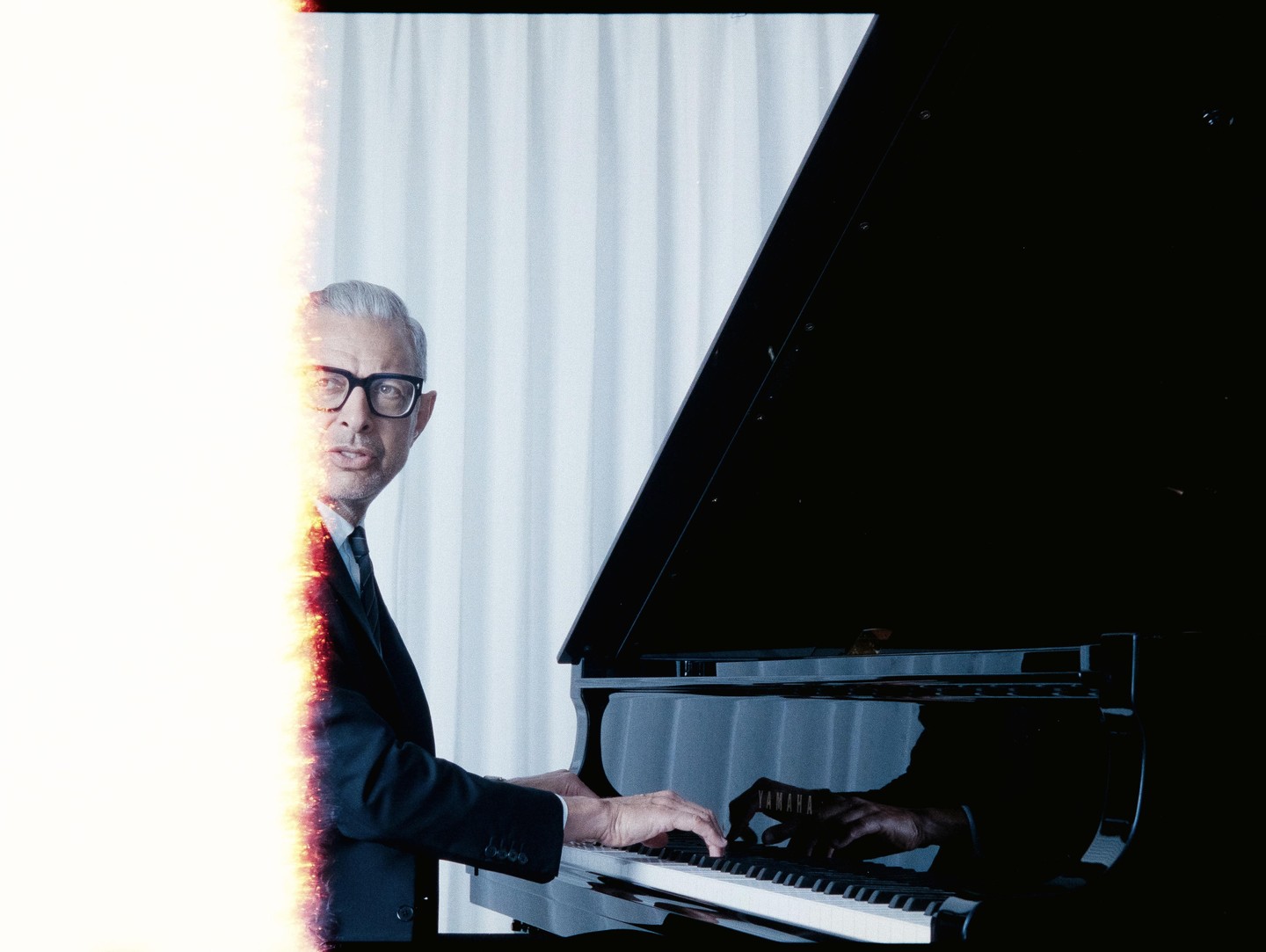 A man in a suit and glasses plays a grand piano, with a bright light leak on the left side of the image.