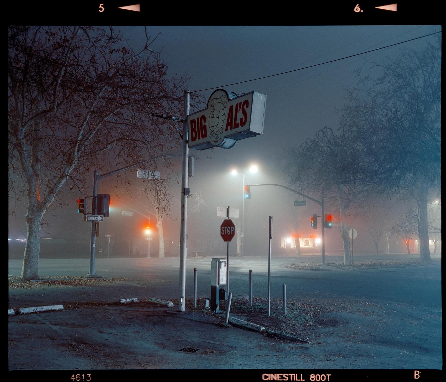A street corner with a "Big Al's" sign and traffic lights is shrouded in fog at night.