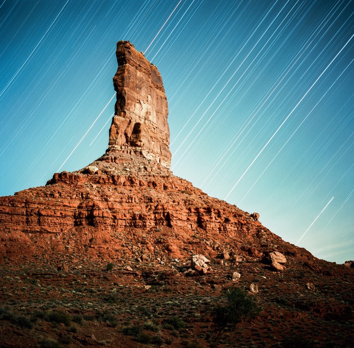 A tall, red rock formation stands against a blue sky with star trails.