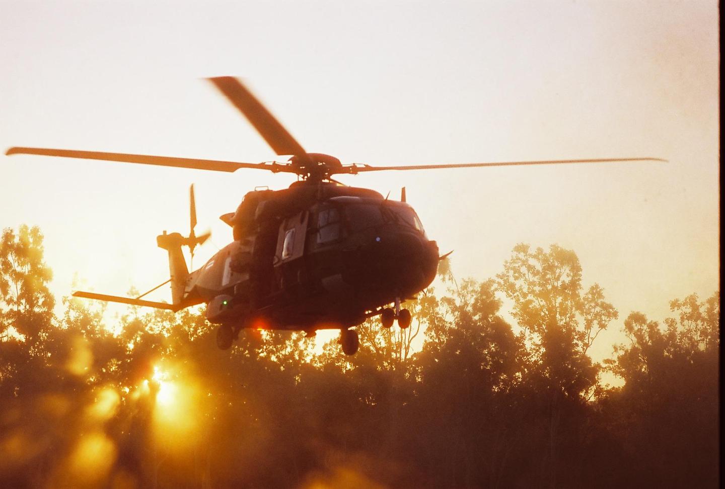 A helicopter is flying over trees with the sun setting behind it.