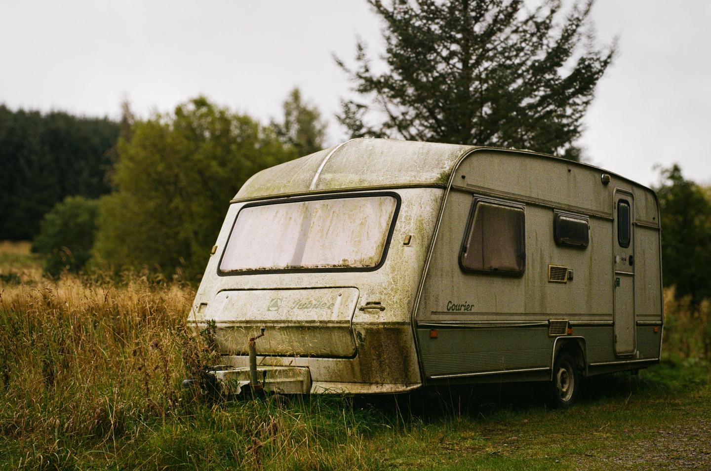 An old, dirty caravan is parked in a field of tall grass with trees in the background.