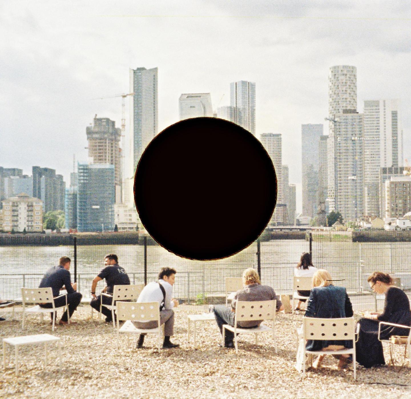 A group of people sit on chairs on a gravel patch next to a body of water, with a city skyline in the background and a large black circle obscuring part of the sky.