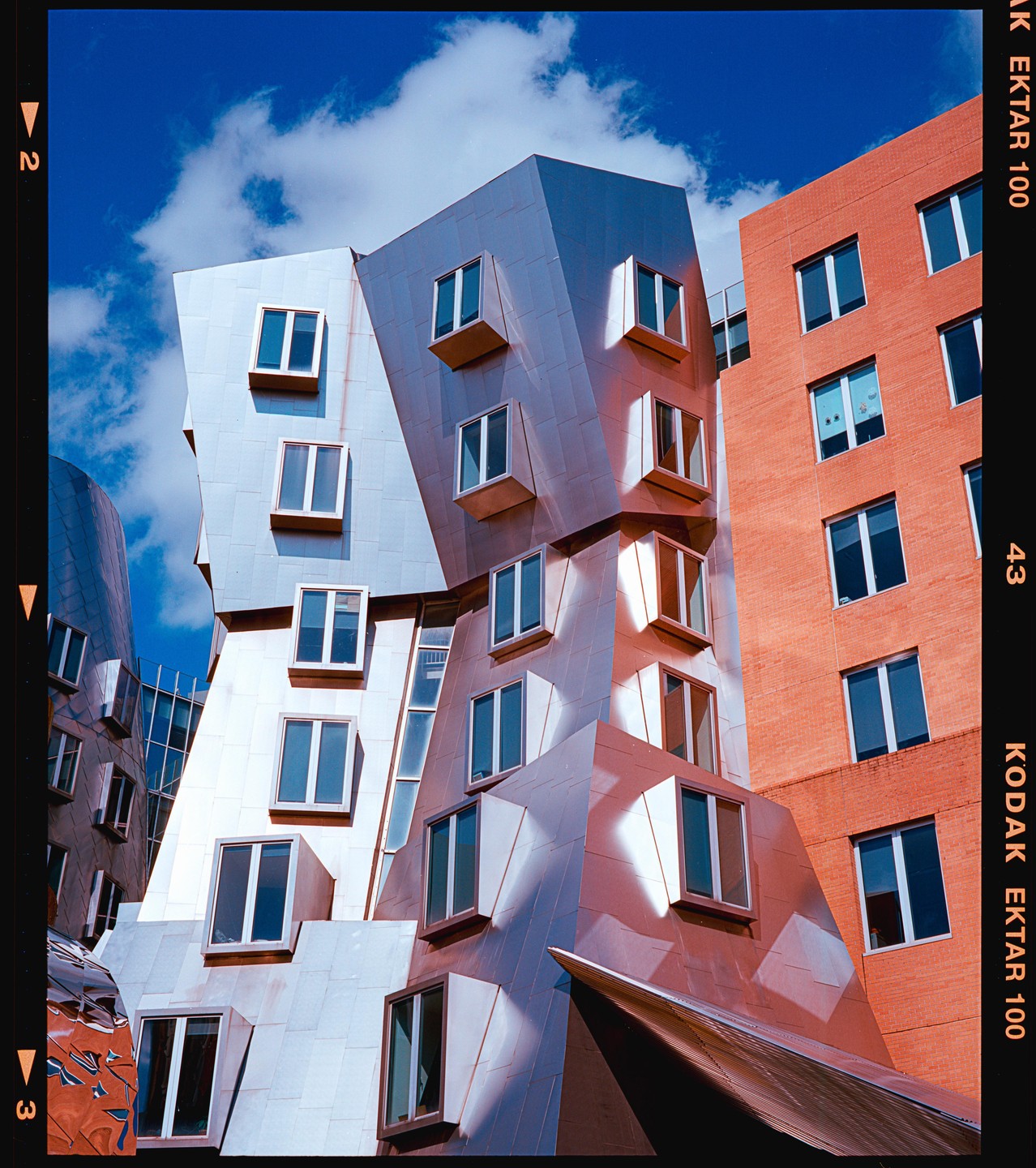 Two buildings with many windows are shown, one with a metallic facade and the other with a brick facade, under a blue sky with clouds.
