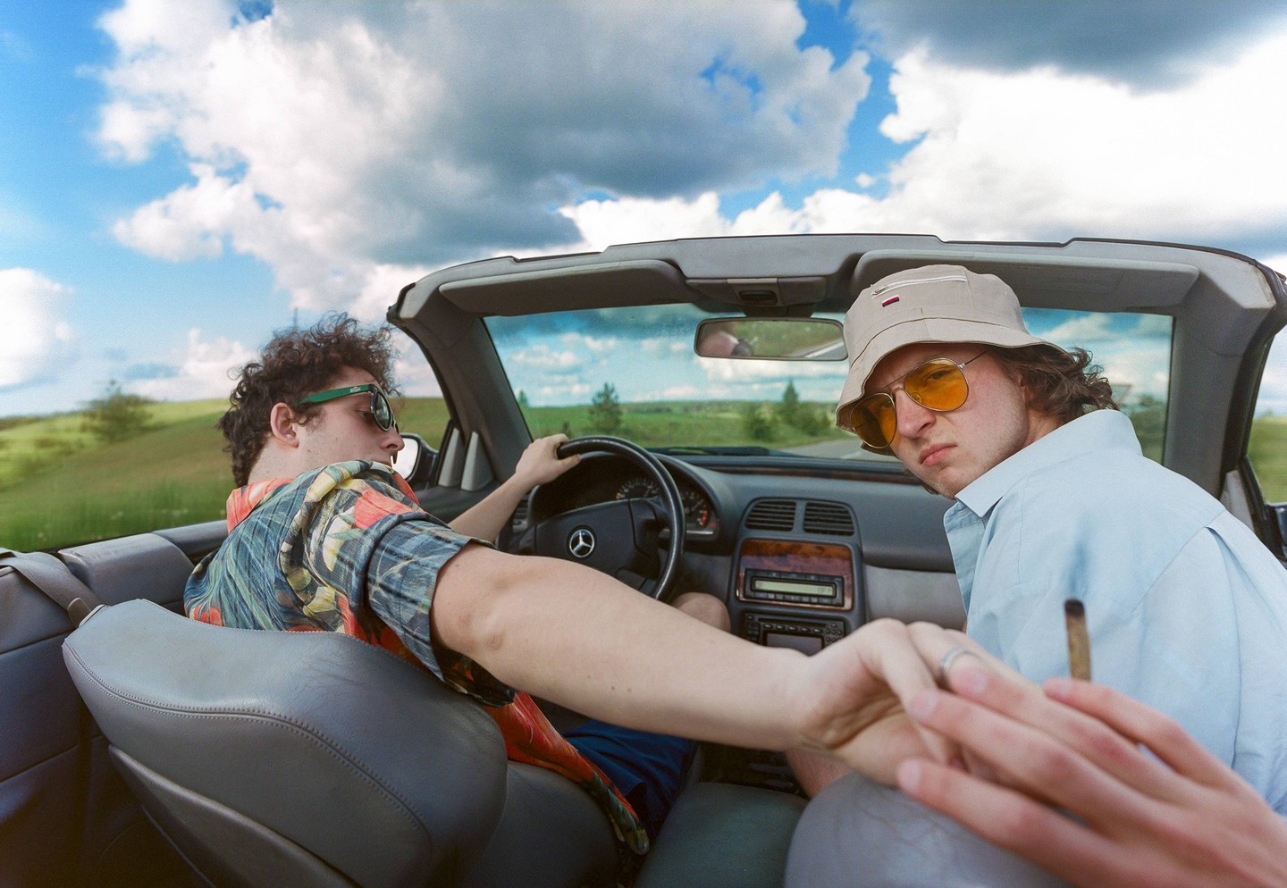 Two men are in a convertible car, one driving and the other looking back at the viewer while holding a joint.