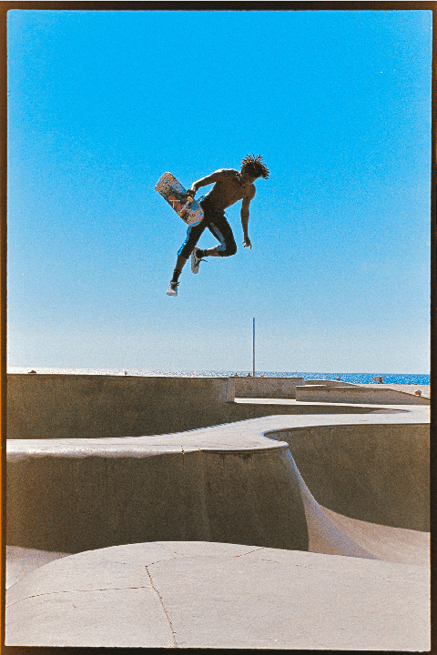 A man with a skateboard is captured mid-air above a concrete skatepark with the ocean visible in the background.