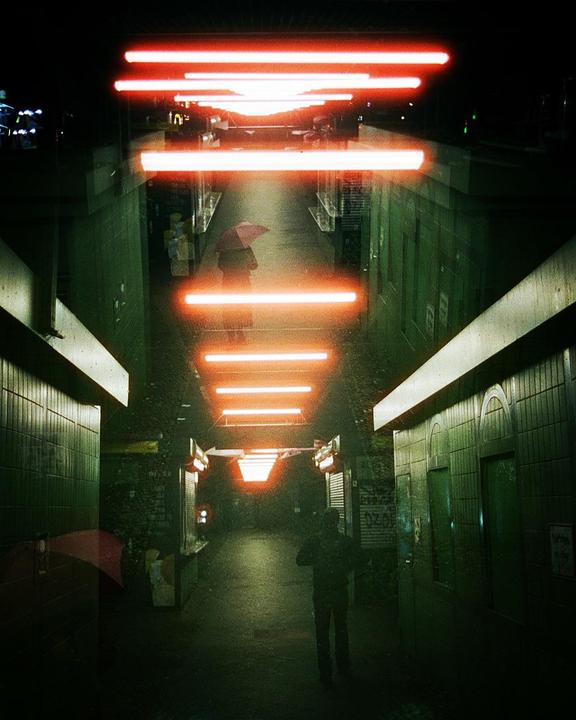 A person with an umbrella walks down a dark, tiled underpass illuminated by bright orange neon lights.