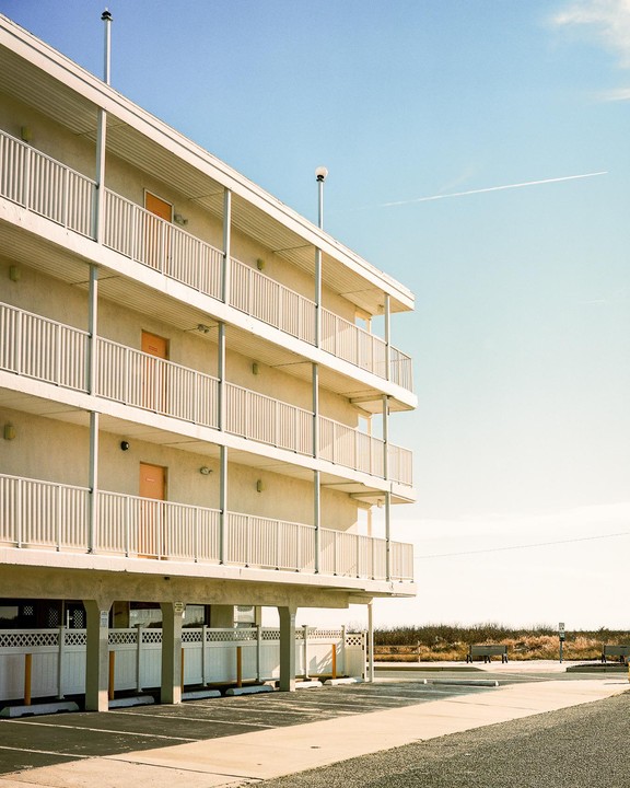 A multi-story motel with balconies and orange doors stands next to a parking lot under a clear sky.