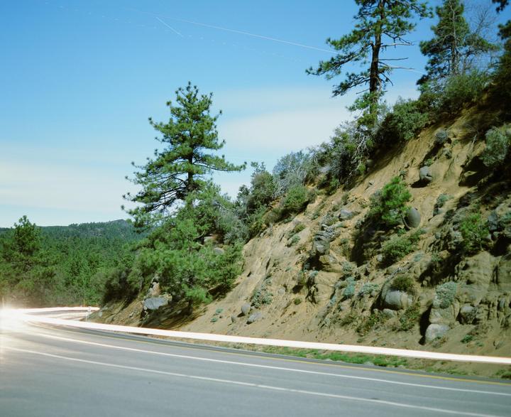 A long exposure photograph captures light trails from vehicles on a winding road next to a steep, tree-covered hillside under a clear sky.