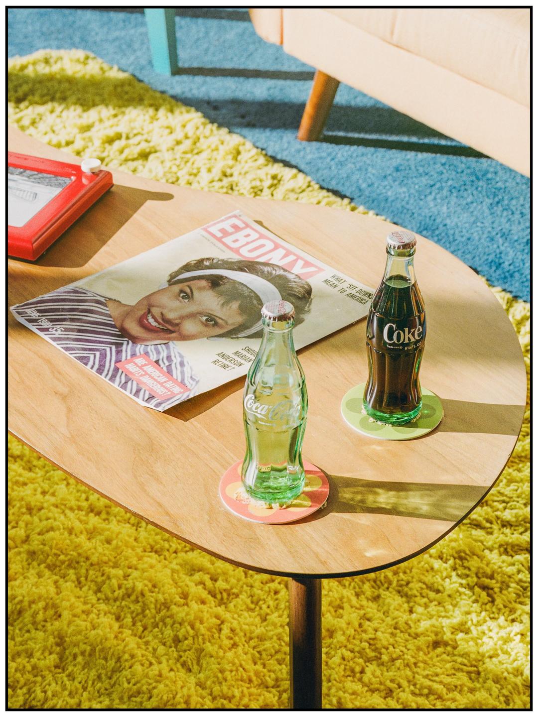 Two glass bottles of Coca-Cola and an Ebony magazine are on a wooden coffee table next to a yellow shag rug.