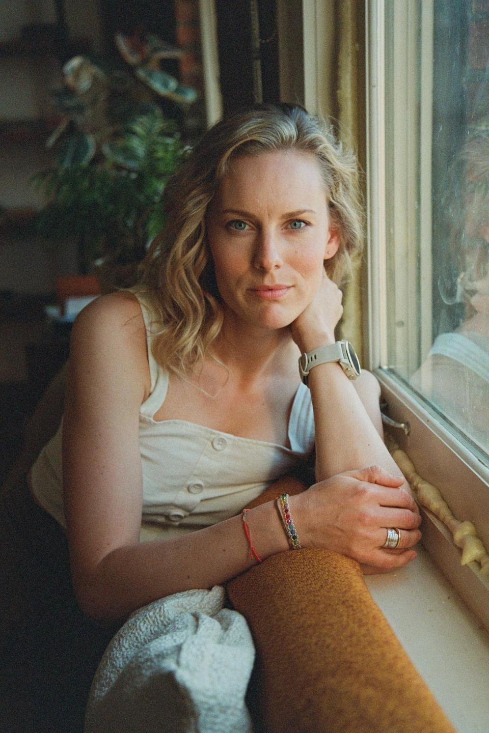 A woman with blonde hair and blue eyes looks directly at the viewer while resting her arm on a window sill.