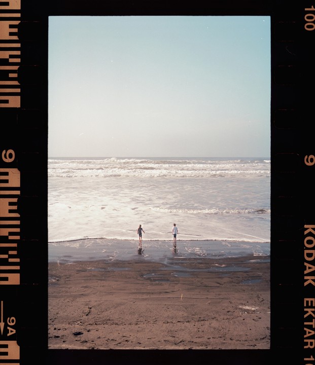 Two children stand in the shallow water at the edge of a beach with waves breaking in the background.