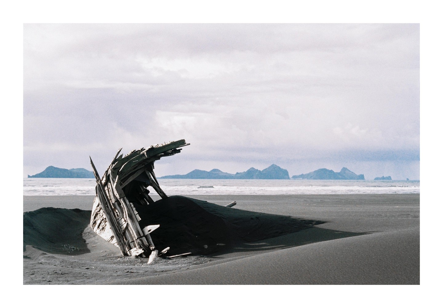 A shipwreck is partially buried in dark sand on a beach with mountains in the distance.