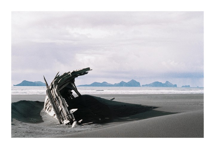 A shipwreck is partially buried in dark sand on a beach with mountains in the distance.