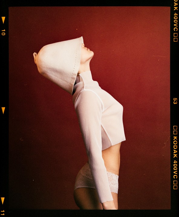 A woman in a white top and underwear with a white hood looks up against a red background.