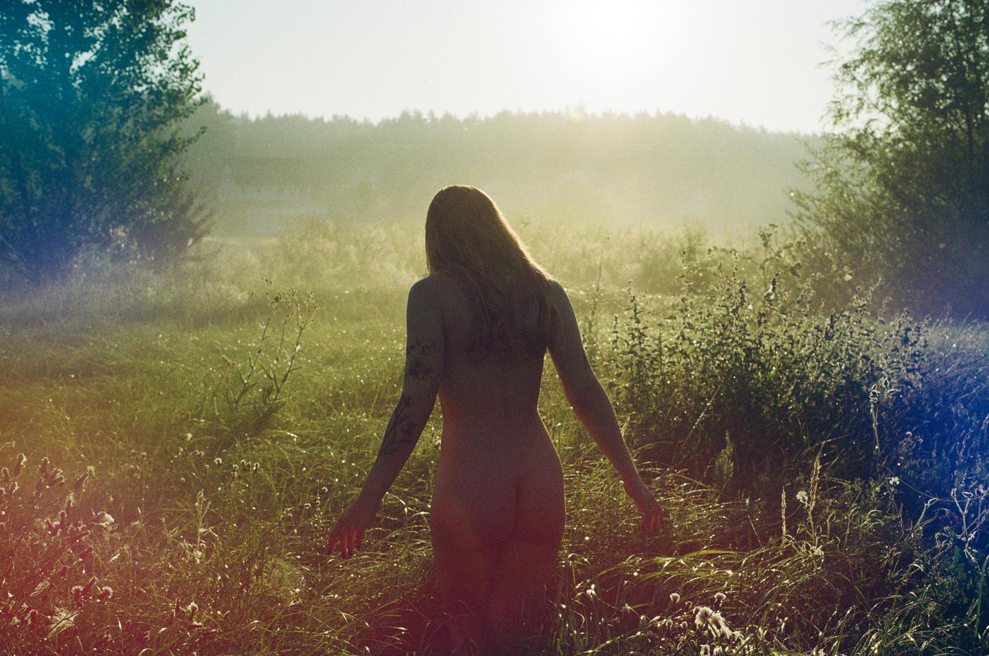 A nude woman with long hair stands in a field of tall grass with trees in the background.