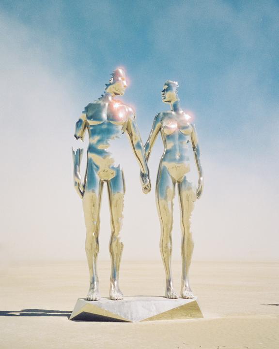 Two metallic, partially broken human figures stand hand in hand on a pedestal in a desert landscape under a bright sky.