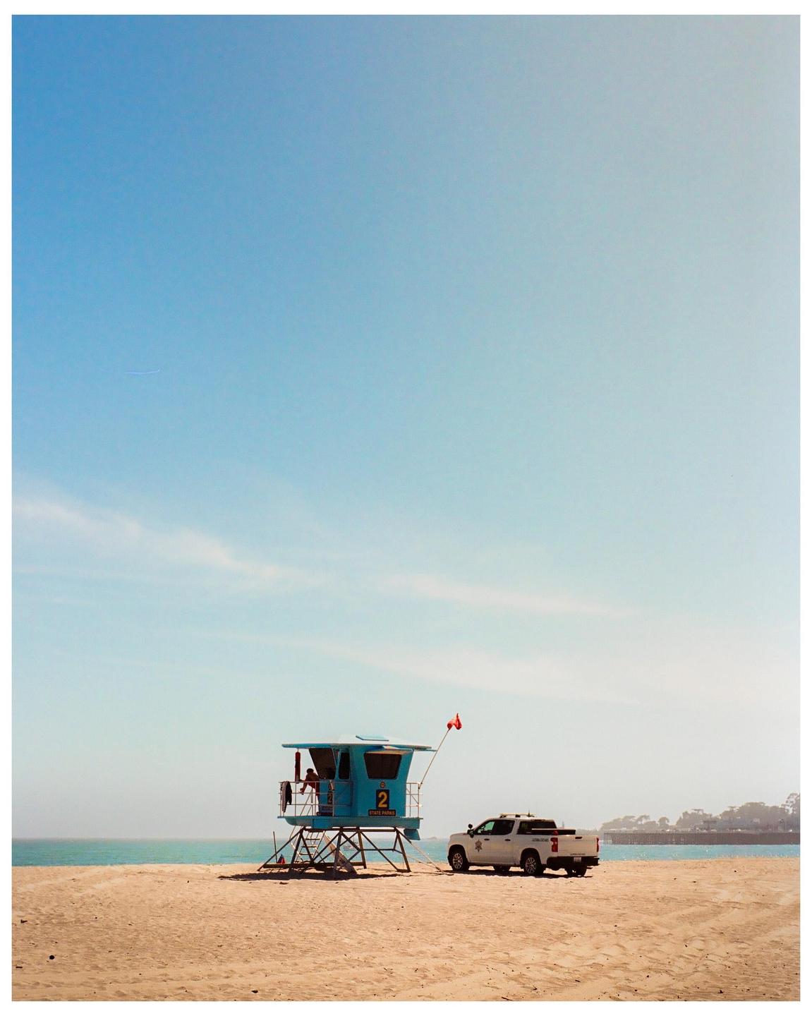 A lifeguard tower and a white truck are parked on a sandy beach next to the ocean under a clear sky.