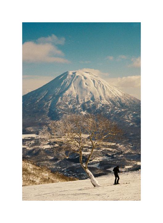 A skier stands on a snowy slope with a large snow-covered mountain in the background.