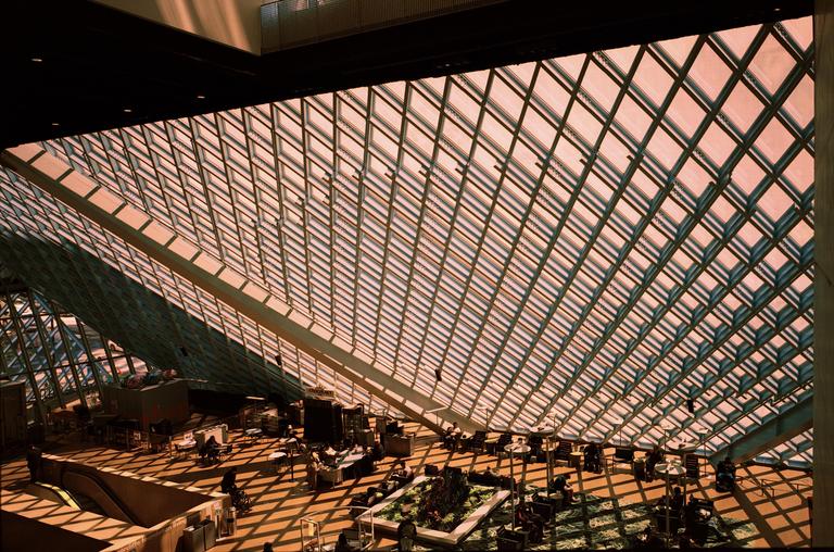 The interior of a modern building features a large glass ceiling and people seated at tables on the floor below.
