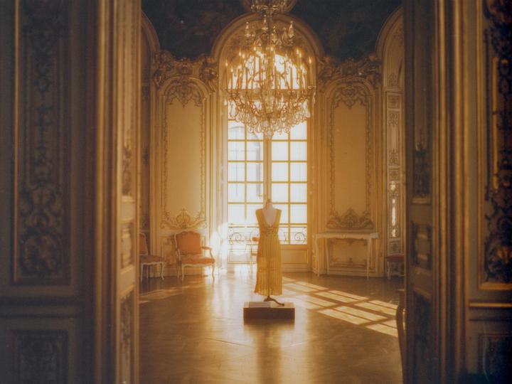 A yellow dress on a mannequin stands in a grand room with ornate gold and white walls, illuminated by sunlight streaming through a large window.