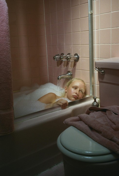A girl with blonde hair is in a bubble bath, looking towards the viewer.