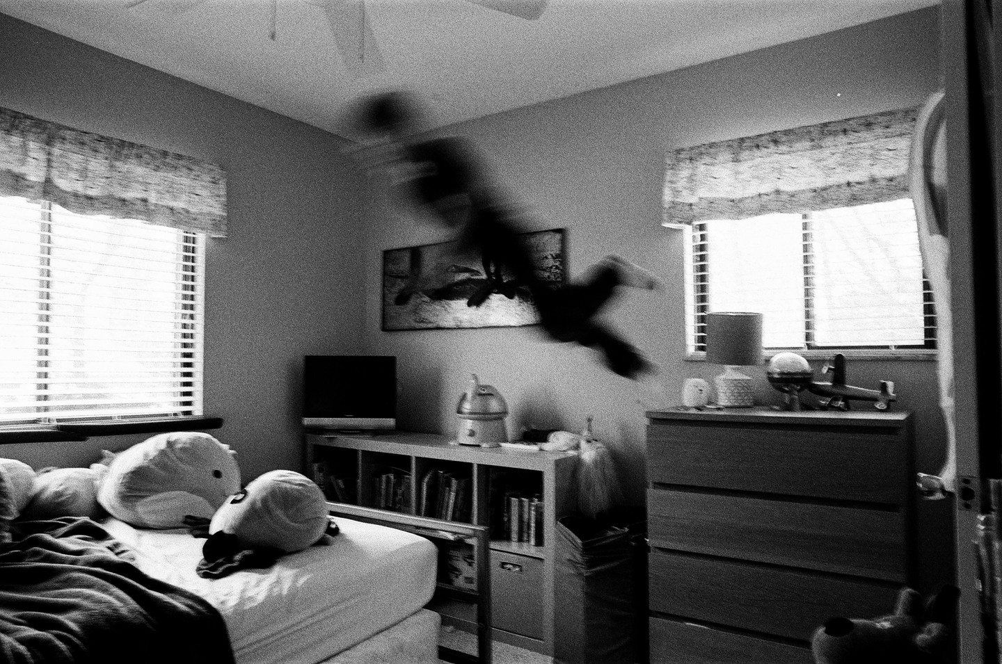 A child jumps in a bedroom with two windows and a bed.