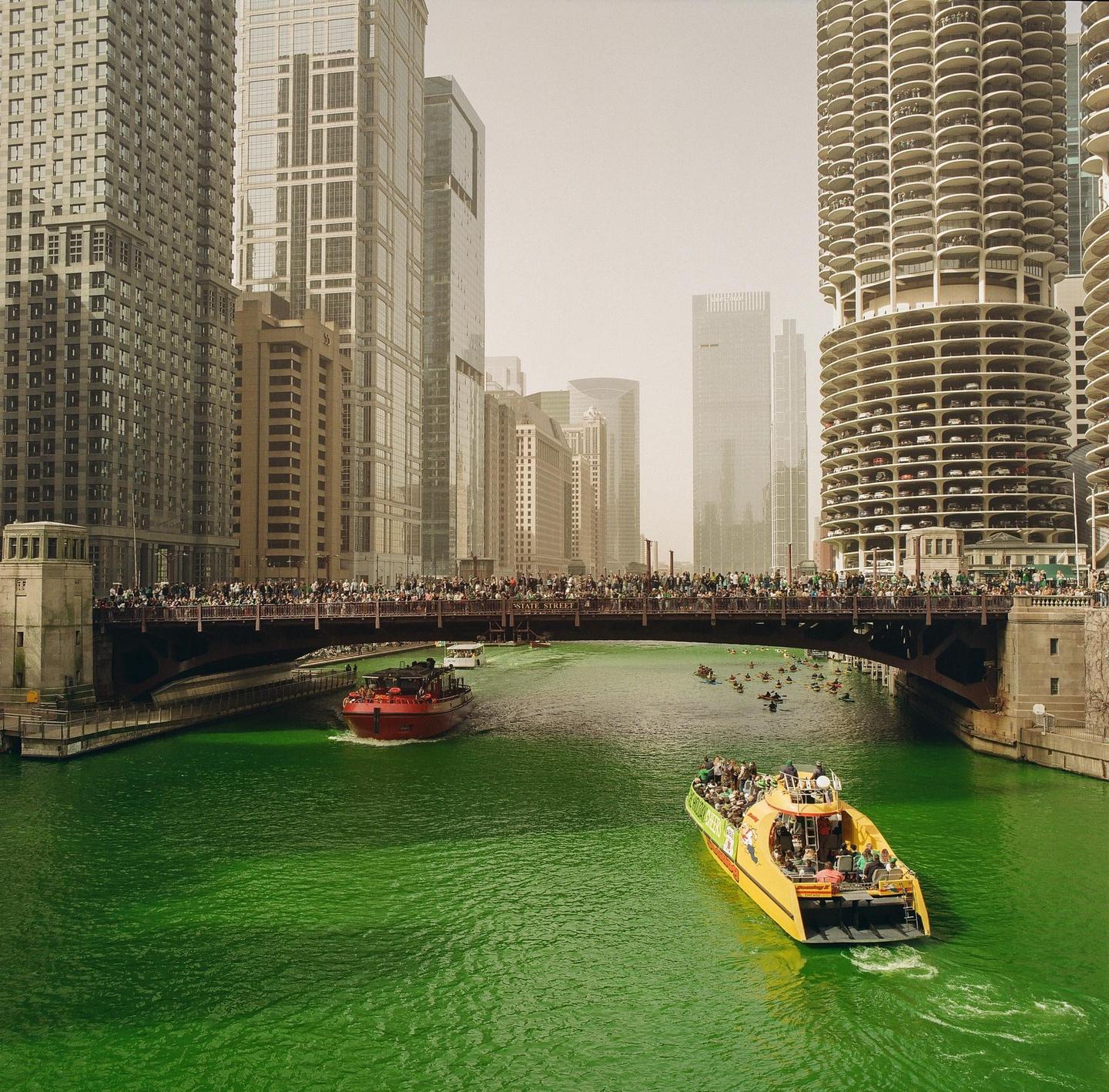 A bridge full of people crosses a bright green river with boats and tall buildings under a hazy sky.
