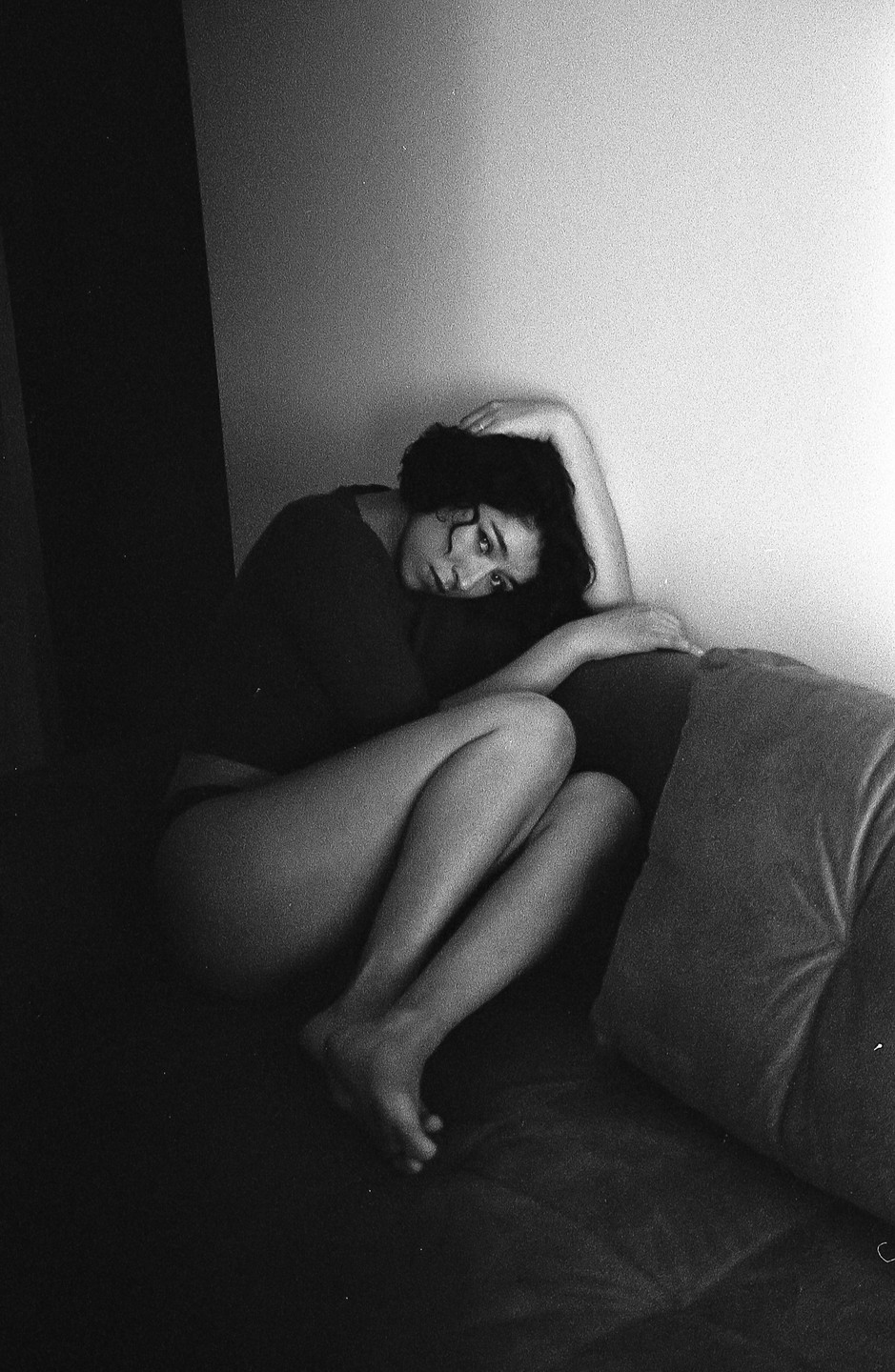 A woman with curly hair is sitting on a couch with her legs bent, looking towards the viewer.