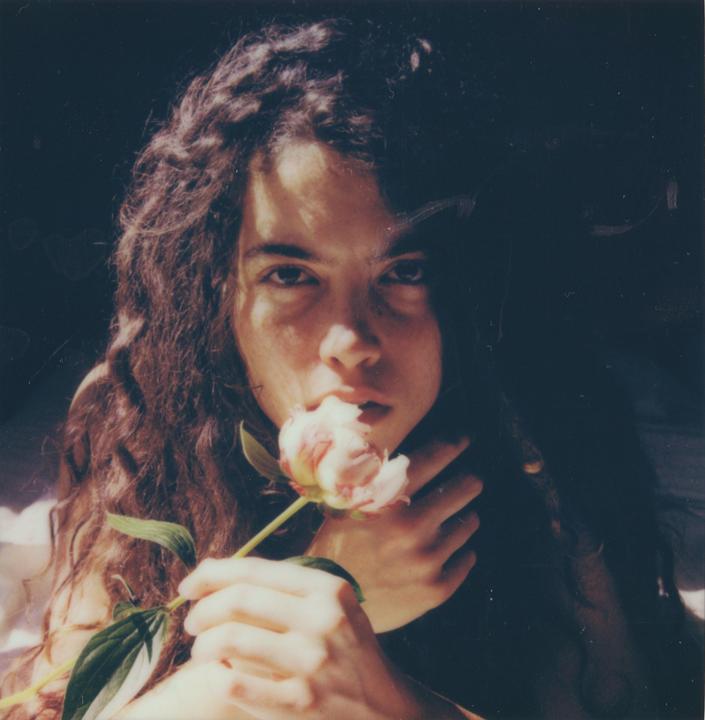 A person with long, dark, curly hair holds a light pink peony to their mouth.