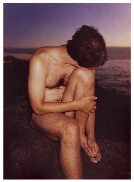A man sits with his head bowed and arms wrapped around his knees by the water at dusk.
