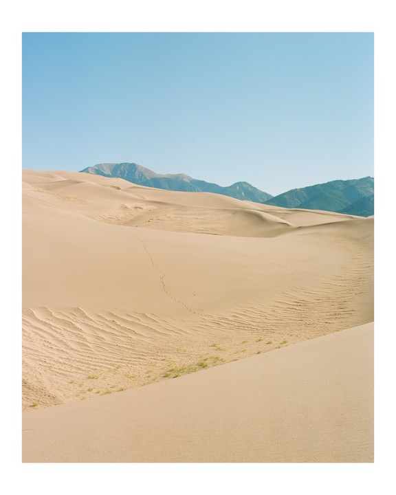 A vast desert landscape features sand dunes with faint footprints and distant mountains under a clear sky.