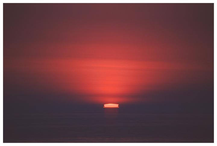 The sun dips below the horizon, casting a red glow across the sky and sea.