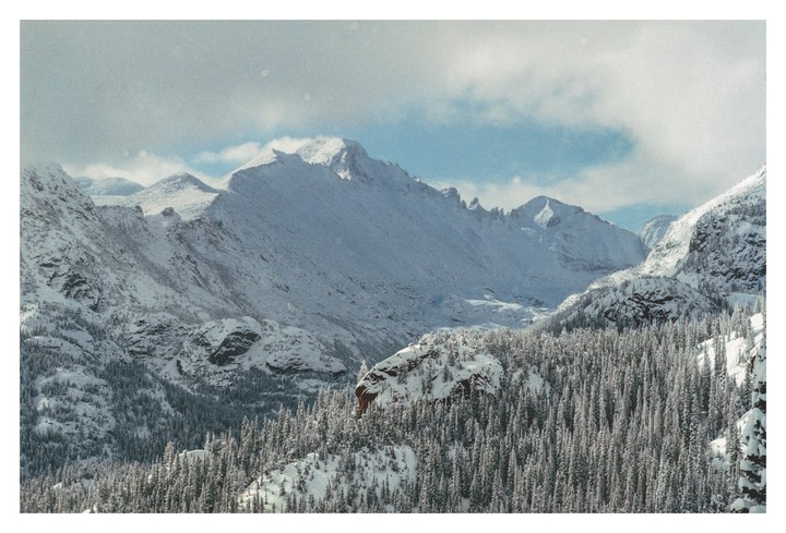 Snow-covered mountains rise above a forest of pine trees under a partly cloudy sky.