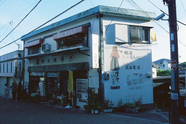 A street corner building with shops on the ground floor and apartments above has a mural of a sake bottle and three children on one wall.