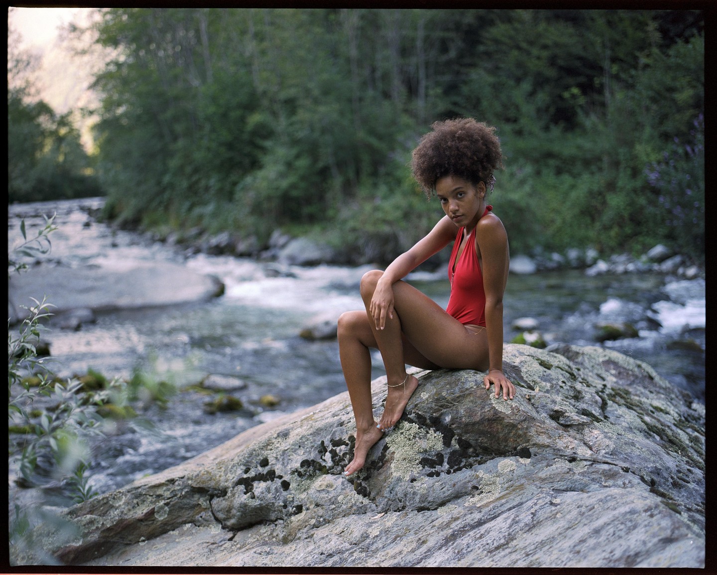 A woman in a red swimsuit sits on a large rock next to a flowing river with trees in the background.