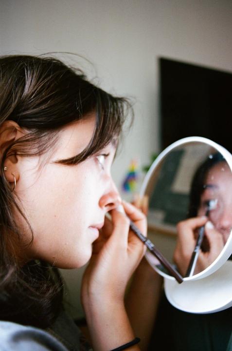 A woman applies eyeliner while looking into a small round mirror.