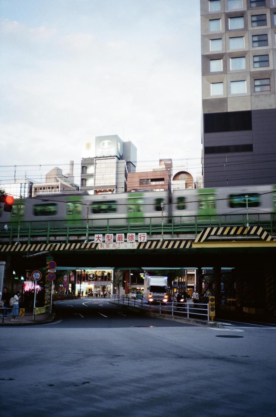 A train passes over a bridge above a street with buildings and vehicles.