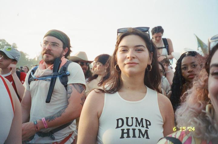 A woman in a white tank top with "DUMP HIM" written on it stands in a crowd of people.