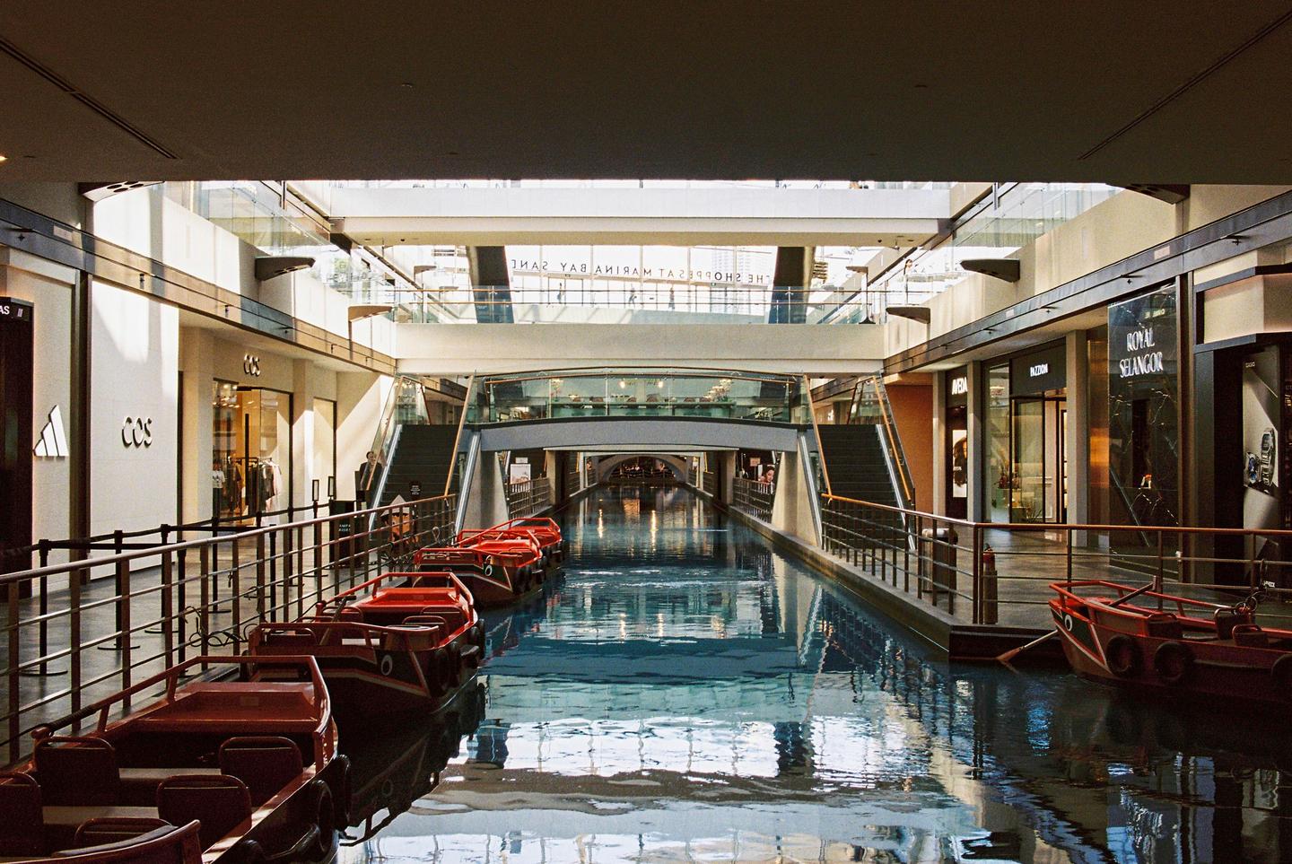A canal with red boats runs through a modern shopping mall with multiple levels and glass railings.