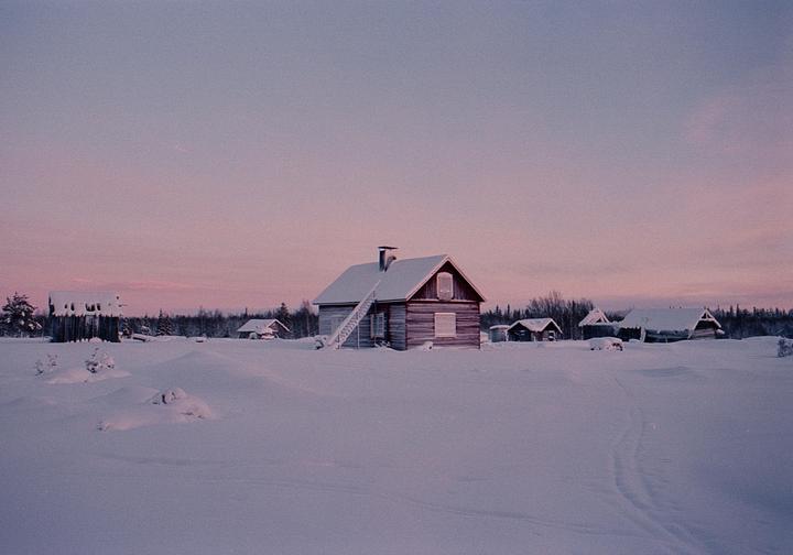 A small wooden house with a snow-covered roof stands in a vast snowy landscape with other small buildings and trees in the distance under a pink and blue sky.
