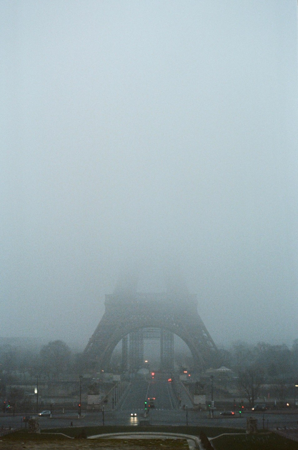 The Eiffel Tower is shrouded in thick fog, with only its base and the surrounding street visible.