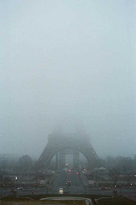 The Eiffel Tower is shrouded in thick fog, with only its base and the surrounding street visible.