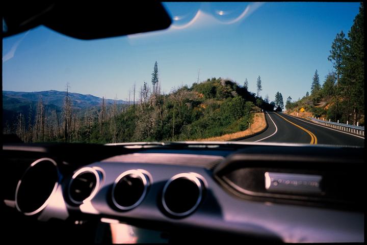 A winding road leads through a mountainous landscape with many trees, as seen from inside a car.