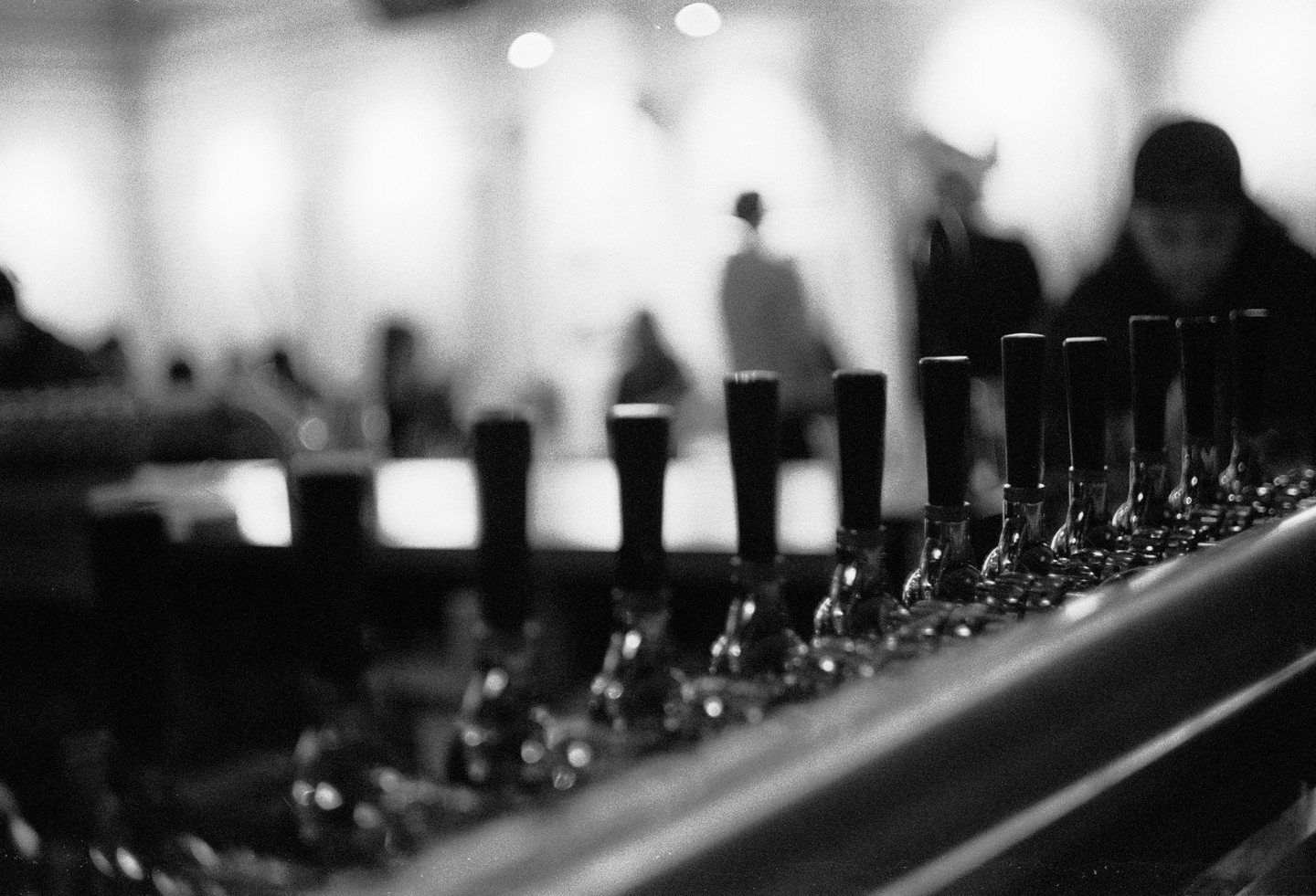 A row of beer taps is in focus in the foreground, with blurry figures of people and lights in the background.