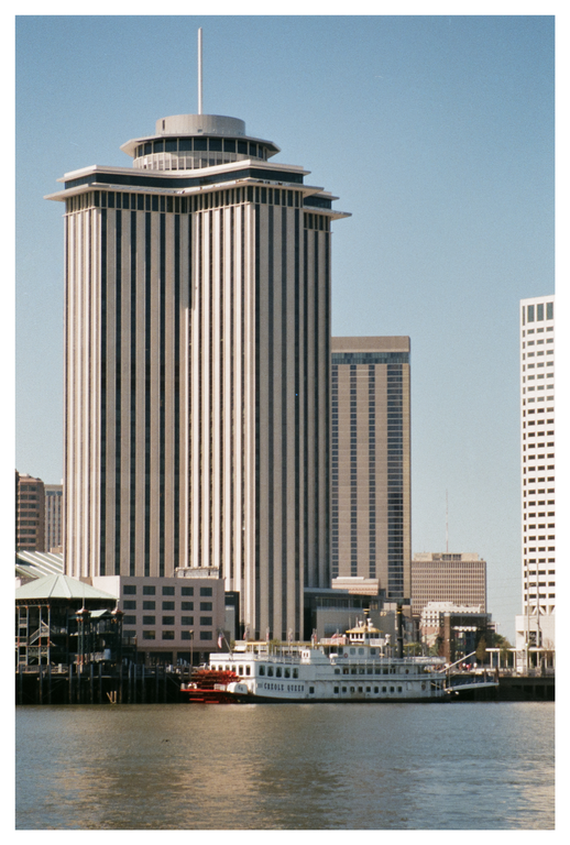 A tall, striped building with a pointed top stands prominently over a river, with a paddleboat docked nearby and other buildings in the background.